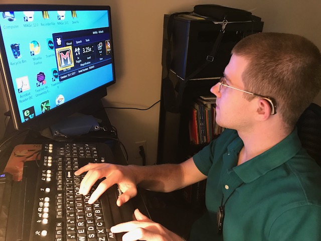A young adult man who is deaf-blind is sitting at a computer with a large monitor screen. He is wearing glasses and hearing aids.