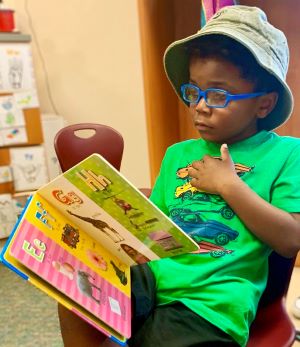 A young boy in a classroom reading a children's book. He is wearing glasses and a hat.