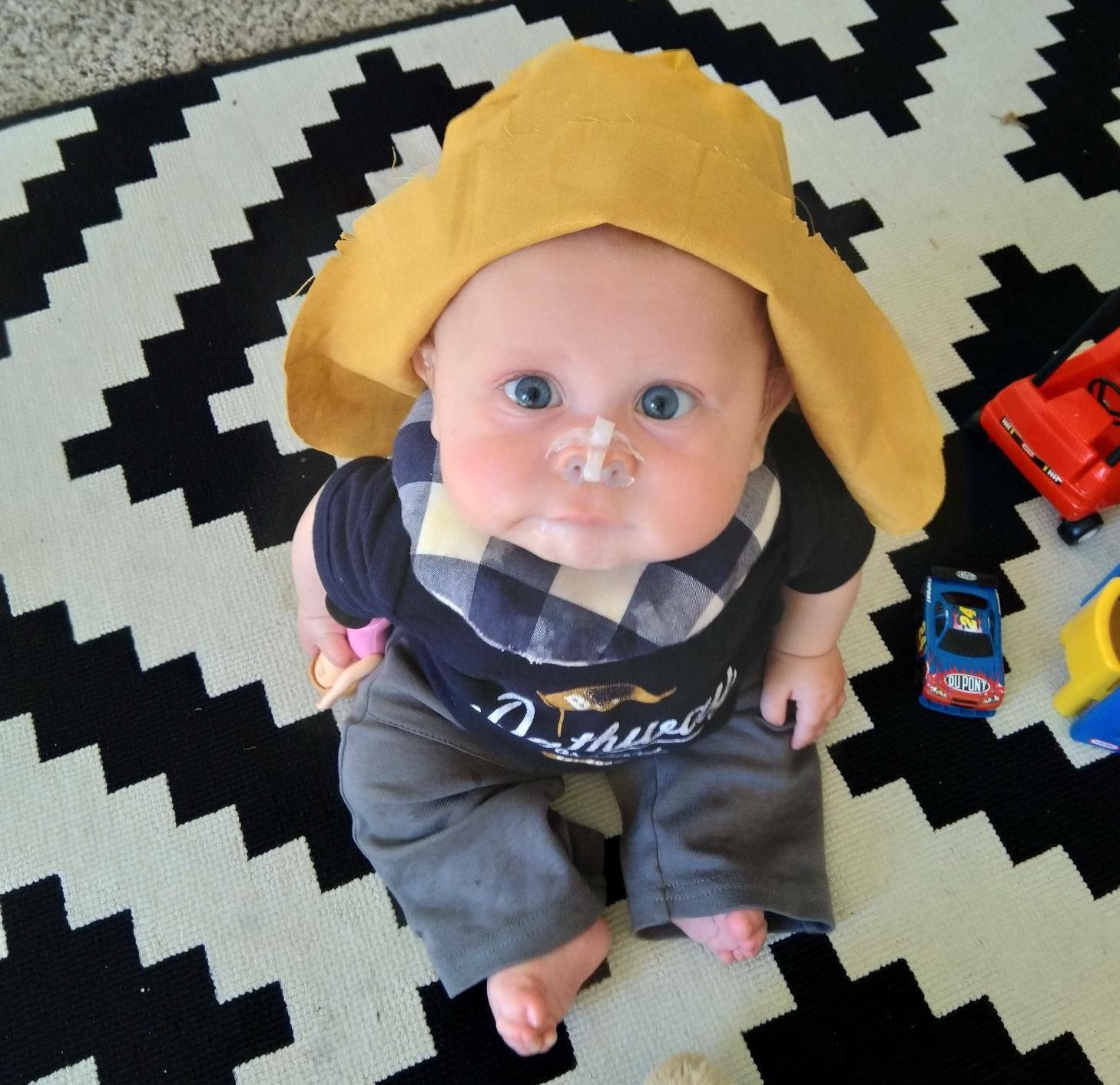 A baby who is deafblind sits on a blanket and looks up toward the camera. He wears a hat and a small device to keep his nostrils open.