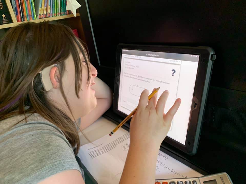 A 13-year-old girl is sitting at her computer smiling. She is touching the screen and holding a pencil in the same hand. She's wearing a hearing aid and her face is very close to the screen.