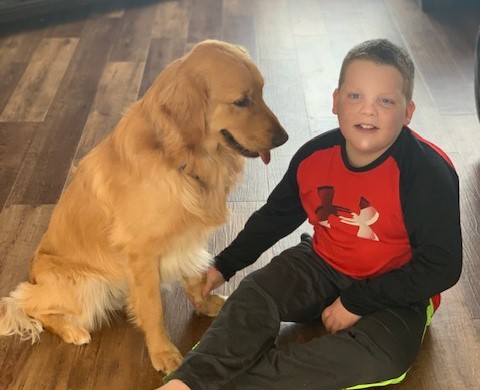 A young boy who is deaf-blind sits on a floor next to his dog, a golden retriever. He is smiling and has his hand on the dogs paw.