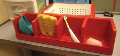 A large red container with four sections, each containing a different object: a bowl, a sponge, a toothbrush, a bar of soap.