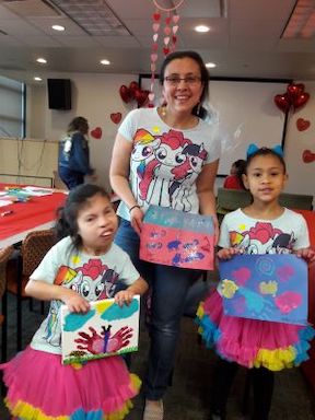 A mother and her two young daughters, one of whom is deaf-blind, stand facing the camera. They are all holding small paintings that they created. In the background is a table with art supplies.